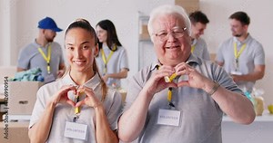 Happy cheerful positive young and senior volunteers doing symbolic heart shape hand sign gesture, smiling and sending you love, support and kind positive feelings. Teamwork in charitable foundation.