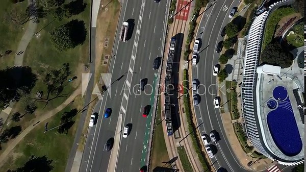Modern day electric tram travels along a city railway passing sporting venues, parks and high-rise recreational areas. Top down drone view