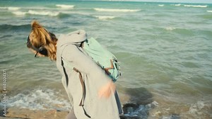 Student wearing headphones with backpack travels along the ocean shore, imagining himself as a bird, making dance moves against the backdrop of sea waves and blue sky.