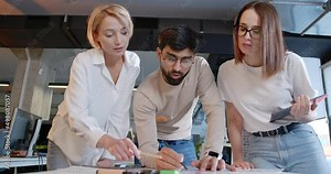 Working team of man and two women standing over the table and charts and discussing project. IT employees. Mixed-races male and females talking and brainstorming. Startup concept.
