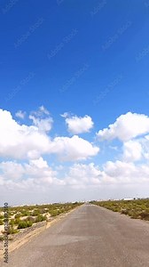 Desert road stretches beneath vibrant blue sky, clouds casting soft shadows over desert terrain, dry landscape blending with open horizon, remote travel adventure desert road