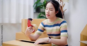 Handheld shot, Young Asian woman wrapping cardboard boxes with duct tape in preparation for moving into new house. Moving house and lifestyle concept.