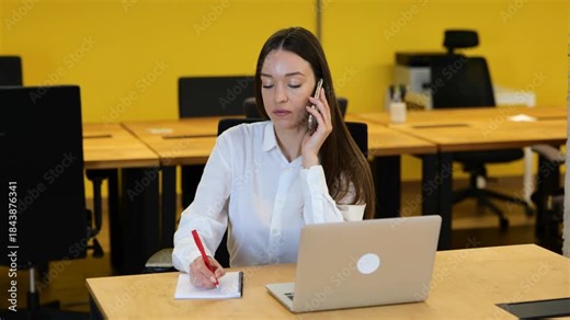 Woman sitting on her desktop, talking by phone and writing down in her notebook