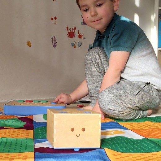 Let me introduce you to our new smiley wooden robot. Cubetto from Primo Toys teaches Alex the basics of programming through adventure and without a screen. Cubetto kit includes a cute robot on wheels, a game board, four types of actions blocks, a mat where the robot can roll around, and an activity book. First Alex places the blocks on the board to tell Cubetto to go straight, turn right or turn left. Once he hits the blue button, the program is transferred wirelessly to Cubetto and it starts mo