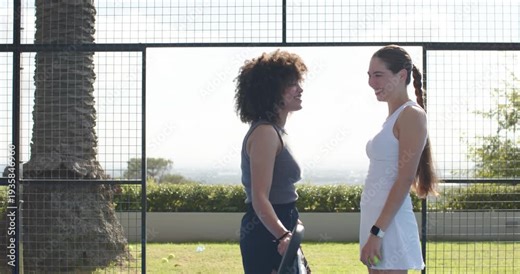 Two adult women standing at low net left pointing, right laughing warming with racket and balls