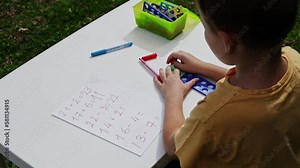 Child with autism has math therapy lesson: uses colorful Numicon shapes, counts, writes answers on paper; sits at white table in sunny garden with bright green grass Stock Video