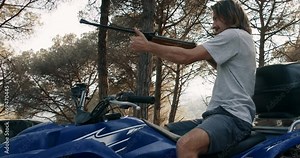 Young man with beard and long hair sitting on quad bike in tho forest on sunset reloads the shotgun and doing fire shoot to the target than going away driving motorcycle