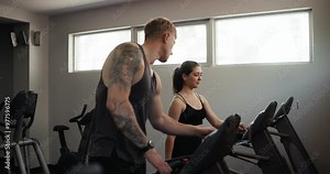 Confident guy trainer athlete with tattoos in a gray T-shirt tells a female client how to properly set up a treadmill for a comfortable workout in the gym