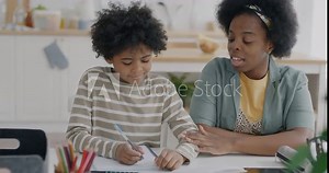African American boy doing homework studying at home while mother helping him with education in kitchen at home. Family and homeschooling concept.