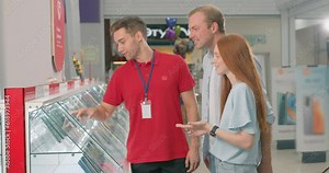 handsome shop assistant in red T-shirt pointing at shelf with latest gadget, showing new model of phone to customers in store of household appliances, electronics and gadgets.
