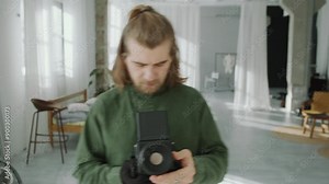 Male photographer with long hair and beard wearing glove looking at camera, telling how to pose, finger-counting and capturing image with professional camera during photo shoot in studio