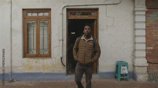 Emerging from doorway man in brown jacket stepping onto sidewalk at worn building, backpack visible