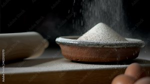 Flour being sieved into a rustic bowl. Baking ingredients and equipment included in frame. Muted colour palette. Slow motion falling flour against a dark background.