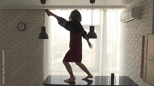 A pretty brunette girl in a velvet dress is dancing barefoot on the black kitchen table with a cellphone in her hand and a wireless speaker on the table