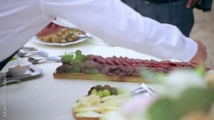 Waiter dressed in uniform serving guests at luxury wedding party outdoors, servant putting salami slices on plate adding cheese chops and other tasty appetizers. Catering service of high level on