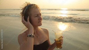 Young playful blonde girl in a swimsuit and large headphones goes along the beach at sunset and dancing to the music with a glass of fruit juice in her hand. Stabilised following camera.