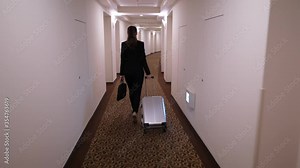 Businesswoman walk to room along hotel passageway, dolly camera follow behind. Woman wear black jacket, carry laptop bag and small trolley case. Overnight stay during assignment, check-in at hotel