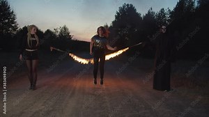Lovely redhead young woman jumping burning skipping rope on dirt road at night, holded by friends in halloween costumes dressed as grim reaper and female vampire during halloween celebration.