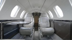 Contemporary interior of private jet cabin. Four white leather seats with headrests arranged in pairs on either side of narrow aisle. Light grey carpet and walls