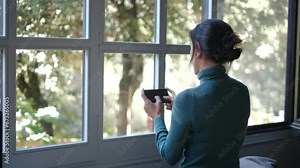 A woman uses her mobile phone to send messages in front of a sunny window. Embracing technology, mobile apps, connecting with people online on a sunny day indoors