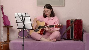 Young woman strumming way to success as classical guitarist, playing six string acoustic guitar with strumming style. Female guitarist honing her craft in home. She is dressed in jeans and polo shirt