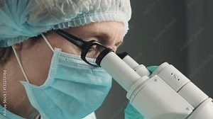 Middle age female scientist in uniform and medical face mask doing microscopic examination in laboratory.