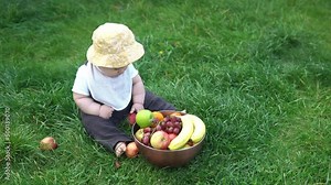 Small Newborn child in summer panama hat sit on grass barefoot in bib with big bowl of fresh fruit. Infant Toddler Boy Taste bites licks apples banana Grapes Garden ouside Healthy eating food harvest