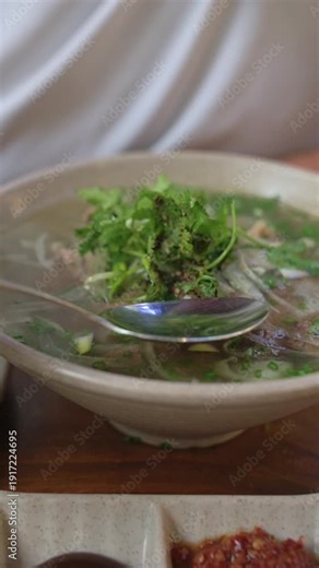 Vertical Screen Bowl Of Vietnamese Pho With Spoon Lifted For Tasting, Caucasian Hand Sampling Clear Broth, Tender Beef Slices And Herb Pile Visible, Cozy Dining Setting With Warm Light