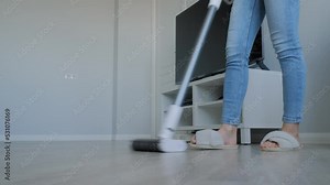 Unrecognizable woman cleaning living room with modern white bagless upright vacuum cleaner - low angle ground view. Technology, housework, routine and housekeeping concept