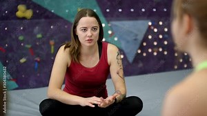 Over-shoulder shot of Caucasian female climbing coach sitting with crossed legs on padded mats next to indoor wall and giving list of instructions to group of trainees