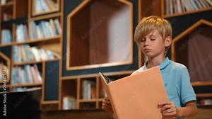 Attractive caucasian clever boy reading a book at library with stack of books placed. Smart child focus on studying, learning from novel or textbook while turning a page. Self-education. Erudition.