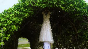 White wedding gown hangs on a wooden hanger from a arbor or passageway of climbing plants 60fps