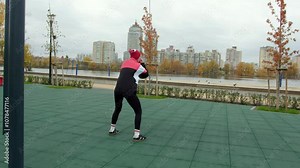 A woman in a red beanie and sportswear performs dynamic squats on an outdoor gym platform, set against an urban riverside view with high-rise buildings and autumn trees.