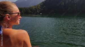 Woman sit on a sunbed in sunglasses and swimming suit. Girl rest on a flood wood underwater pier. The pavement is covered with water in lake. Background are mountain and forest. Swimwear bikini blue.