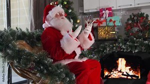 Angry Santa Claus uses phone understands modern technology sitting in his workshop near the fireplace, Christmas tree and holiday gifts.