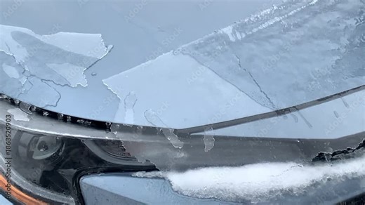 A thick sheet of ice being scraped from the front windshield of a car after freezing rain. Ice buildup from winter weather is removed using an ice scraper, highlighting hazardous driving conditions, f