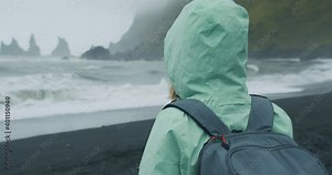 Woman with a green raincoat in mood rainy weather visiting black sand beach looking at Reynisfjara sea stack cliff formations in sea, Iceland.