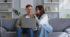Happy young family couple using a laptop computer. Cheerful wife sits down on the sofa next to her husband who is online shopping on his notebook PC and they continue together