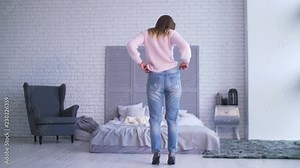 Rear view of gorgeous adult woman wearing old loose ragged jeans, showing her success after loosing weight while standing in bedroom. Attractive slim female in big pants showing successful weight loss