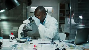 Black forensic scientist examining evidence under microscope and taking notes at desk in crime lab