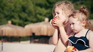 Two little girls eat apples on the beach together. Healthy eating concept