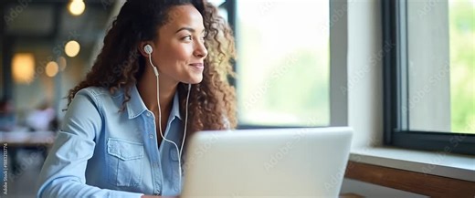 Smiling woman working on a laptop, headphones in, while sunlight streams through office windows; camera slowly pans, and ambient office lights flicker gently for a cinematic, professional atmosphere.