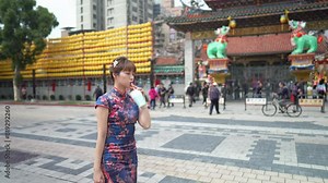 A young Taiwanese woman in a blue traditional cheongsam dress sips bubble tea while walking past historic buildings in Wanhua District, Taipei City, Taiwan. slowmotion