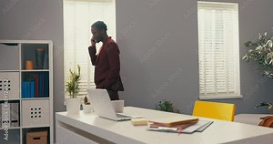 Elegant serious man with dark skin working in high position manager boss walks around room against gray wall by window, dressed in maroon suit talking on the phone, important company call