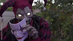 Close-up scary jester Halloween decoration with glowing eyes and a fanged grin. The harlequin holds a white theatrical mask with red and black makeup in a backyard scene.
