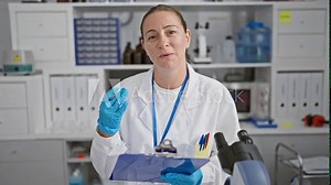 Speaking science! beautiful, young blonde scientist woman caught mid-conversation, clipboard in hand, passionately communicating medical research in bustling laboratory work environment.
