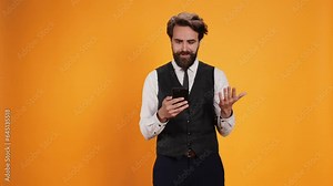 Before serving guests at table, classy butler checks phone looking at online web page against yellow background. Staff member with smartphone serving in an eatery, catering sector.