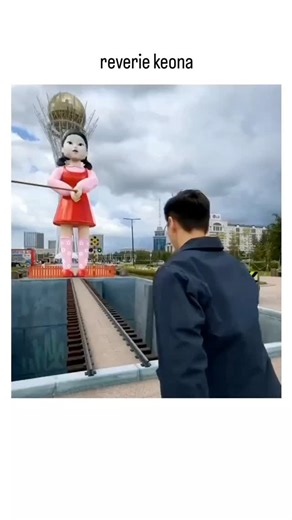 buzz__decode on Instagram: "This post shares a 11-second video clip depicting a man in black sprinting along a precarious elevated track toward the real-life giant Young-hee doll statue from Squid Game, filmed at its original location in Jincheon County, South Korea's carriage museum. The dynamic footage, likely captured via drone for smooth aerial perspectives, creates high tension as the runner navigates barriers and nears the animatronic doll, prompting viewer speculation in Indonesian-langua
