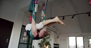 Man hanging upside down on aerial sinks. Pan around tilt down handheld shot of strong male hanging upside down on aerial hammock and bending body during yoga session in light studio
