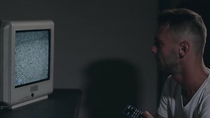 Broken Television or TV Propaganda. Mind Control - Zombified Man in White T-shirt Switches Channels on the Old TV in Dark Room. TV Signal Search. Distortion on the Screen. Medium Close-Up, Film Grain